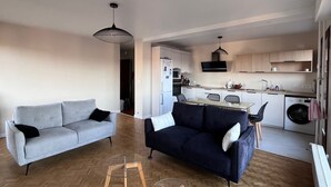 Un espace de vie compact comprenant un canapé gris, un canapé foncé, une table basse en verre et un coin cuisine équipé d’un four à micro-ondes, d’une cuisinière et d’un lave-linge.