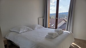 Un lit bien fait, avec du linge de lit et des serviettes blanches, un plancher en bois et une fenêtre donnant sur des immeubles et des collines.