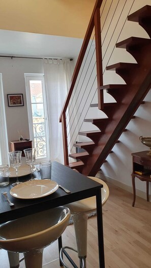 Uma sala de jantar com mesa posta para dois, escada de madeira e janela com cortinas.