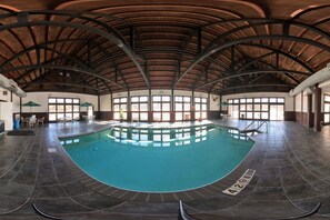 An indoor swimming pool with a curved design, surrounded by a high ceiling with wooden beams, and furnished with tables and chairs. - Coratel Plus Suites by Jasper Black Riverfalls - 2 Queen Beds Non-Smoking (Black Riverfalls)