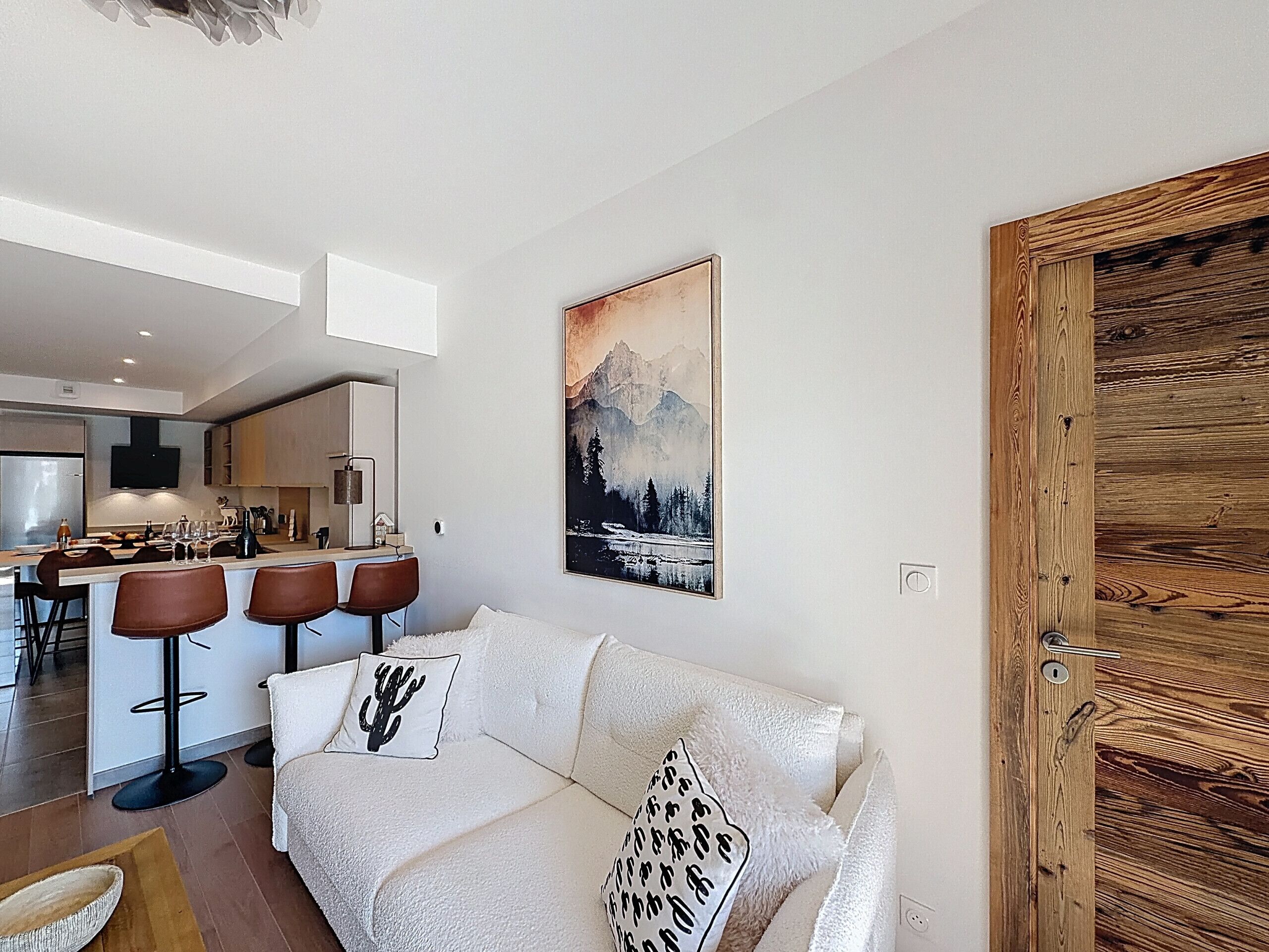 Une cuisine moderne avec un îlot central, un coin repas doté de tabourets de bar, un canapé blanc agrémenté de coussins décoratifs et une porte en bois.