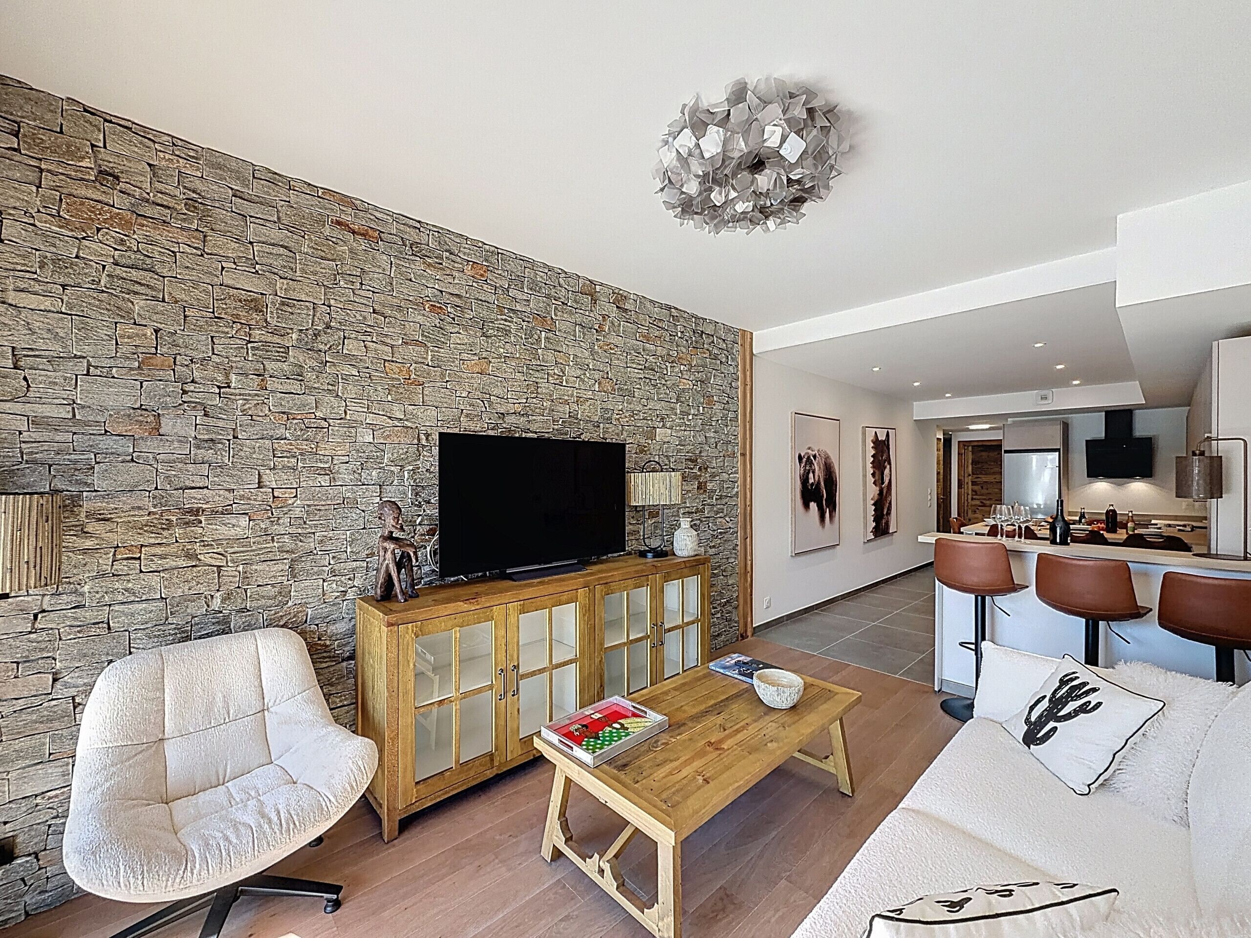 Un salon moderne avec un mur en pierre, un téléviseur, un meuble en bois, un canapé blanc, une table basse et un coin repas avec des tabourets de bar.