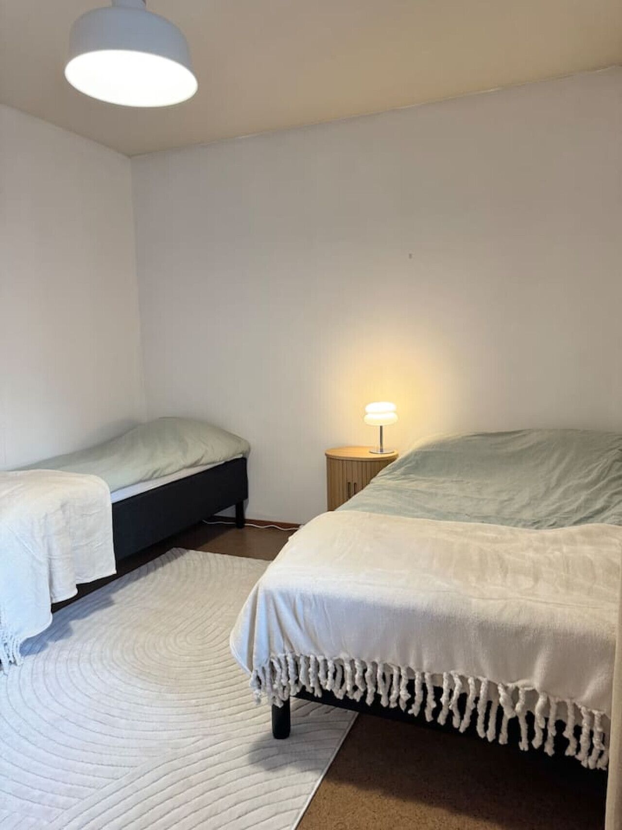 A bedroom with two single beds, a bedside table with a lamp, and a large bed with a white bedspread.