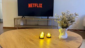 A round wooden table with two lit candles and a vase of flowers, a television displaying the Netflix logo, and a metal stand in front of the TV. - Bright & Spacious Retreat Near Santa Claus Village (Rovaniemi)