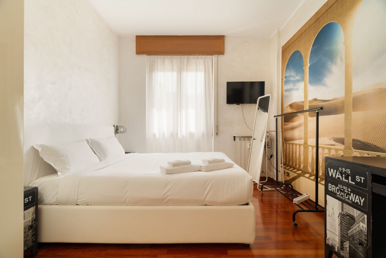 Ein Hotelzimmer mit einem großen Bett, einem Flachbildfernseher, einem Fenster mit durchsichtigen Vorhängen und einer Wandmalerei einer Wüstenszene.