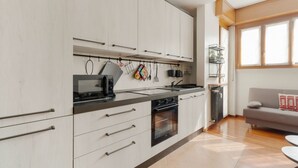 A modern kitchen with white cabinets, a black stove, and a dining area with a grey sofa and a small table. - GuestHost - Comfortable 60m2 apartment, ideal for 4, on the 4th floor of a building with lift (steps to reach it). Located in Fiera Milano City between Portello and CityLife, a historic exhibition area now transformed into the Allianz MiCo convention cen (Milan)
