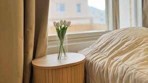 Une chambre à coucher avec un lit, une table de chevet ornée d’un vase de fleurs et une fenêtre donnant sur des immeubles.