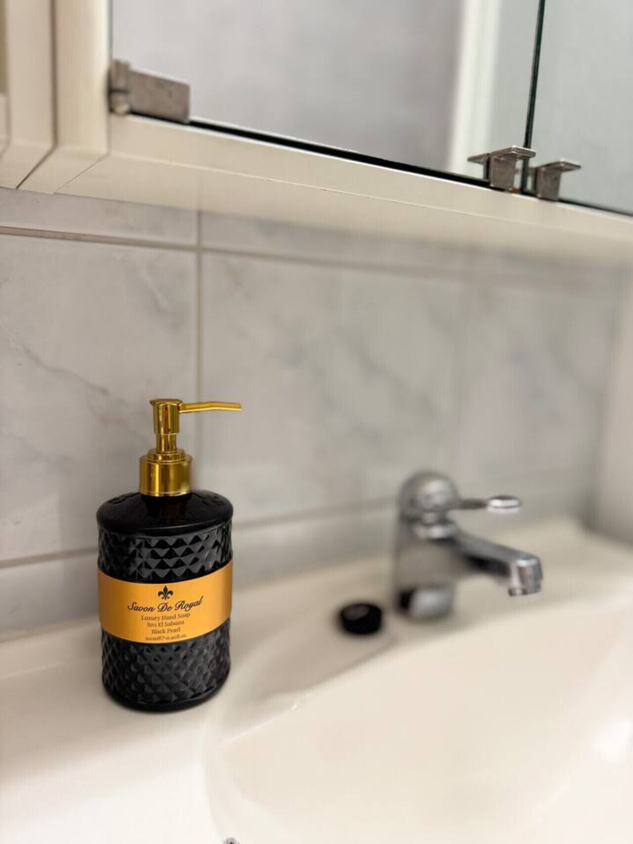 Un bagno con un erogatore di sapone nero etichettato 'Savoir De Royal' e un lavandino bianco con un rubinetto d'argento.