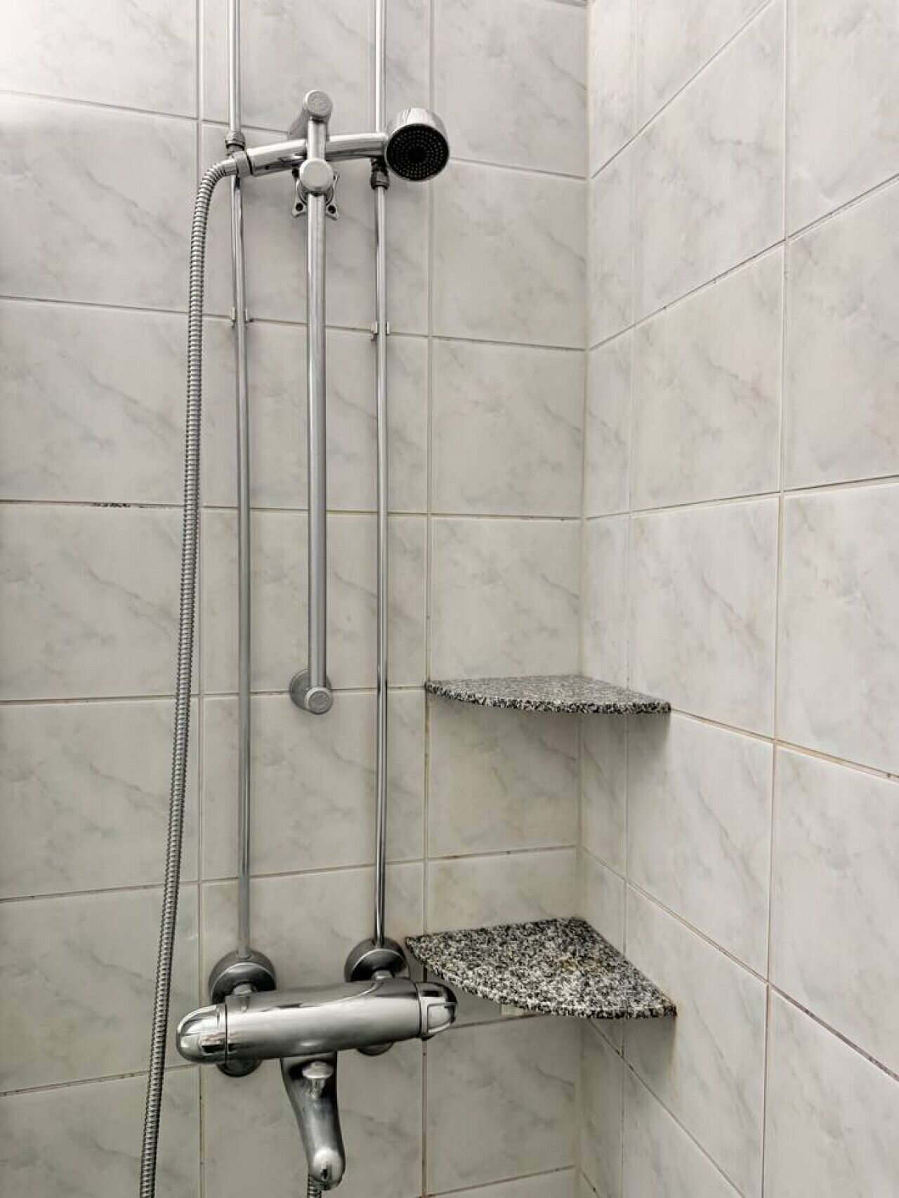 A shower with a handheld showerhead, a wall-mounted faucet, and a corner shelf.