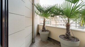 Un balcon agrémenté de deux palmiers en pot, doté d’un sol carrelé et d’une fenêtre donnant sur un immeuble.