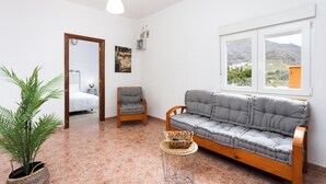 Ein Wohnzimmer mit einer Couch, einem Sessel, einer Topfpflanze und Blick auf eine Berglandschaft durch ein Fenster.