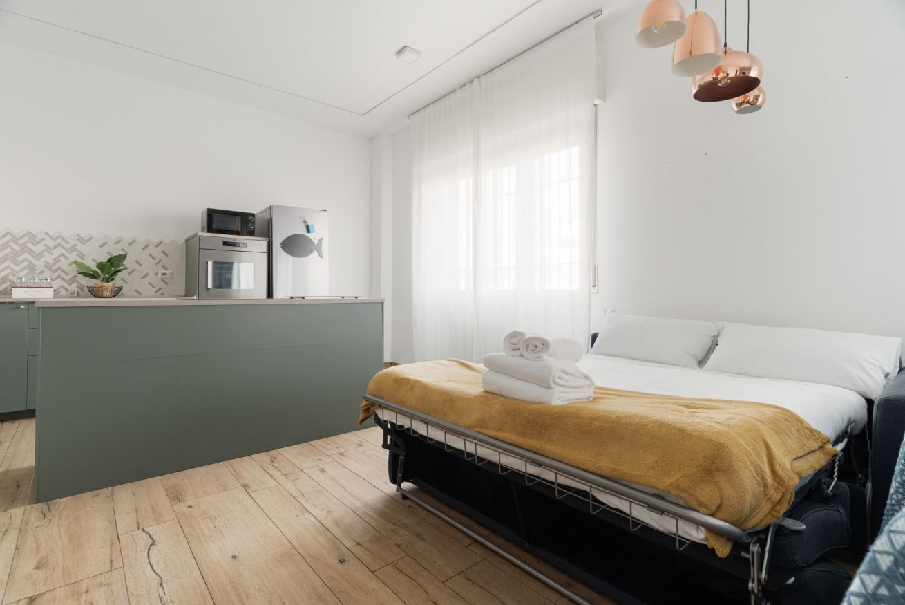 A compact hotel room with a kitchenette, a bed with a yellow blanket, and a window with sheer curtains.