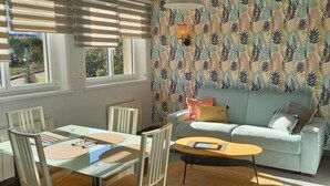 A dining area with a glass table and chairs, a light blue sofa, patterned wallpaper, and a wall-mounted lamp. - 3* Gîte: Port & Beach, 50m from the Sea, South-Facing, Quiet Location (Fécamp)