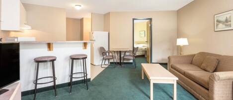 Habitación de hotel con kitchenette, zona de comedor, sala de estar con sofá y televisor, y alfombra verde.