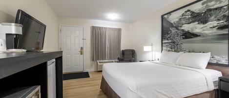 A hotel room with a bed, a desk with a coffee machine, a chair, a lamp, and a large framed picture on the wall.