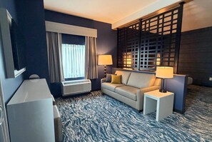 Habitación de hotel con sofá, dos mesitas de noche con lámparas, un ventanal con cortinas, un equipo de aire acondicionado y un separador decorativo.