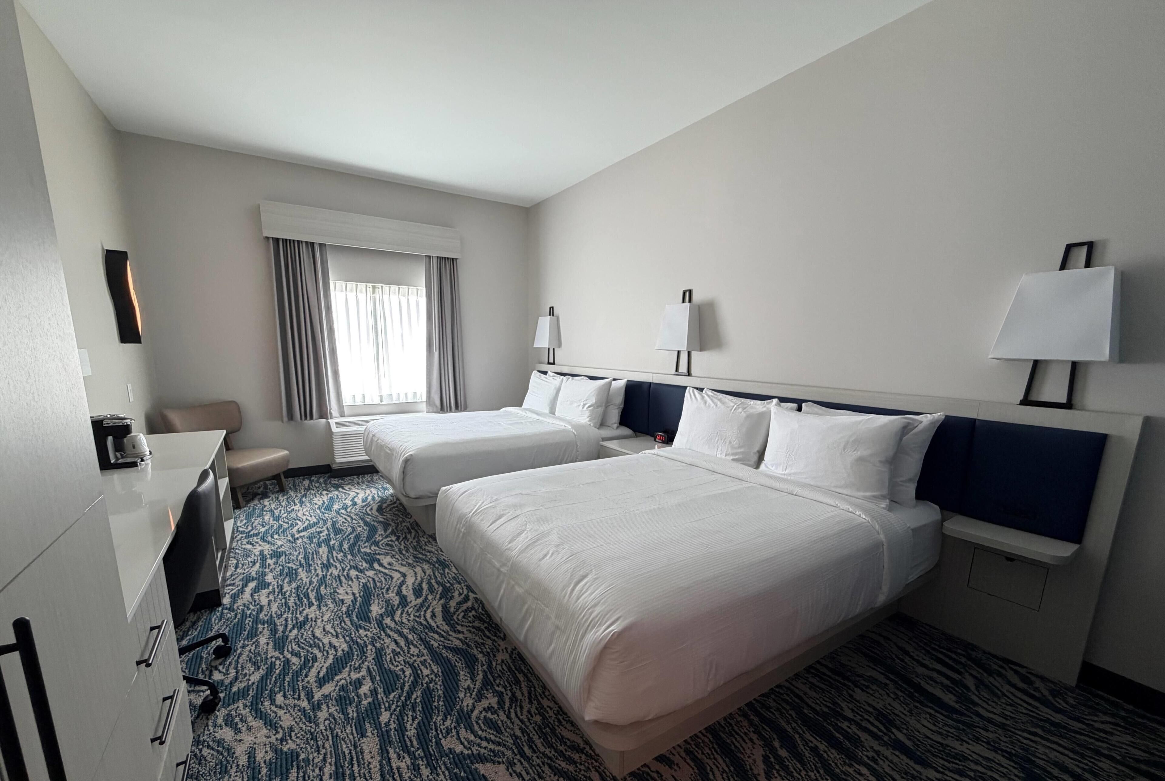 Habitación de hotel con dos camas, un escritorio, una silla y un ventanal con cortinas.
