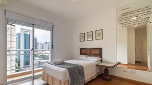Hotelkamer met een bed, nachtkastje, groot raam met uitzicht op de stad en een muur met ingelijste kunstwerken.