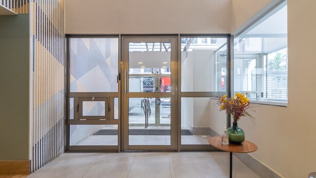 L’entrée d’un hôtel, avec une porte en verre, un sol carrelé et une petite table sur laquelle est posé un vase de fleurs.