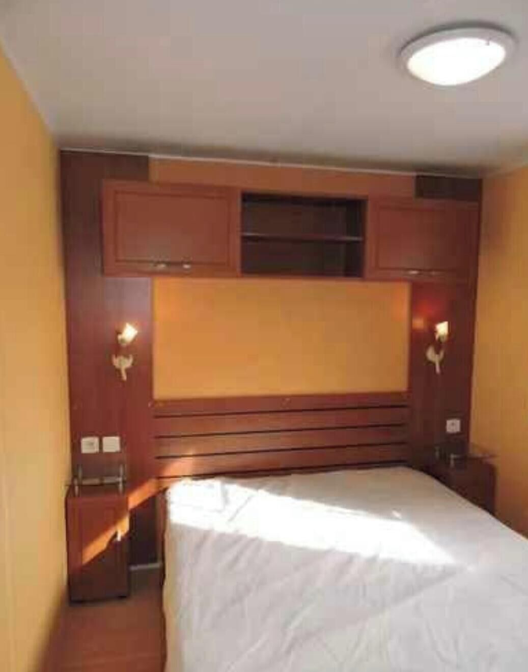 A bedroom with a bed, wooden headboard, wall-mounted light fixtures, and a ceiling light.