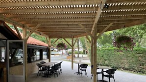 An outdoor patio with wooden tables and chairs, a covered seating area, and hanging flower baskets. - 🌞 Air-conditioned mobile home 6 pers - Camping 3⭐ avec piscine - Landes - dès 70€/nuit (Sainte-Eulalie-en-Born)