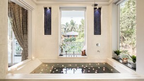 Une salle de spa avec une baignoire, une fenêtre donnant sur des palmiers et une petite table avec des bougies.