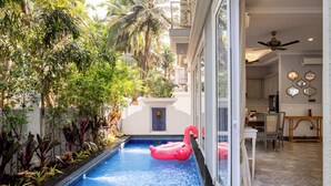 Une piscine agrémentée d’un flotteur en forme de flamant rose, entourée de plantes tropicales et bordée d’un bâtiment blanc au sol carrelé.