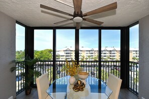 Ein Essbereich mit einem Glastisch, weißen Stühlen, einem Deckenventilator und Blick auf einen Gebäudekomplex durch große Fenster.