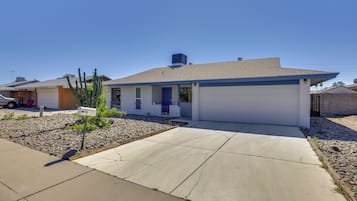 Une maison d’habitation avec un garage, un petit jardin devant la maison agrémenté d’un cactus, et un ciel bleu dégagé.