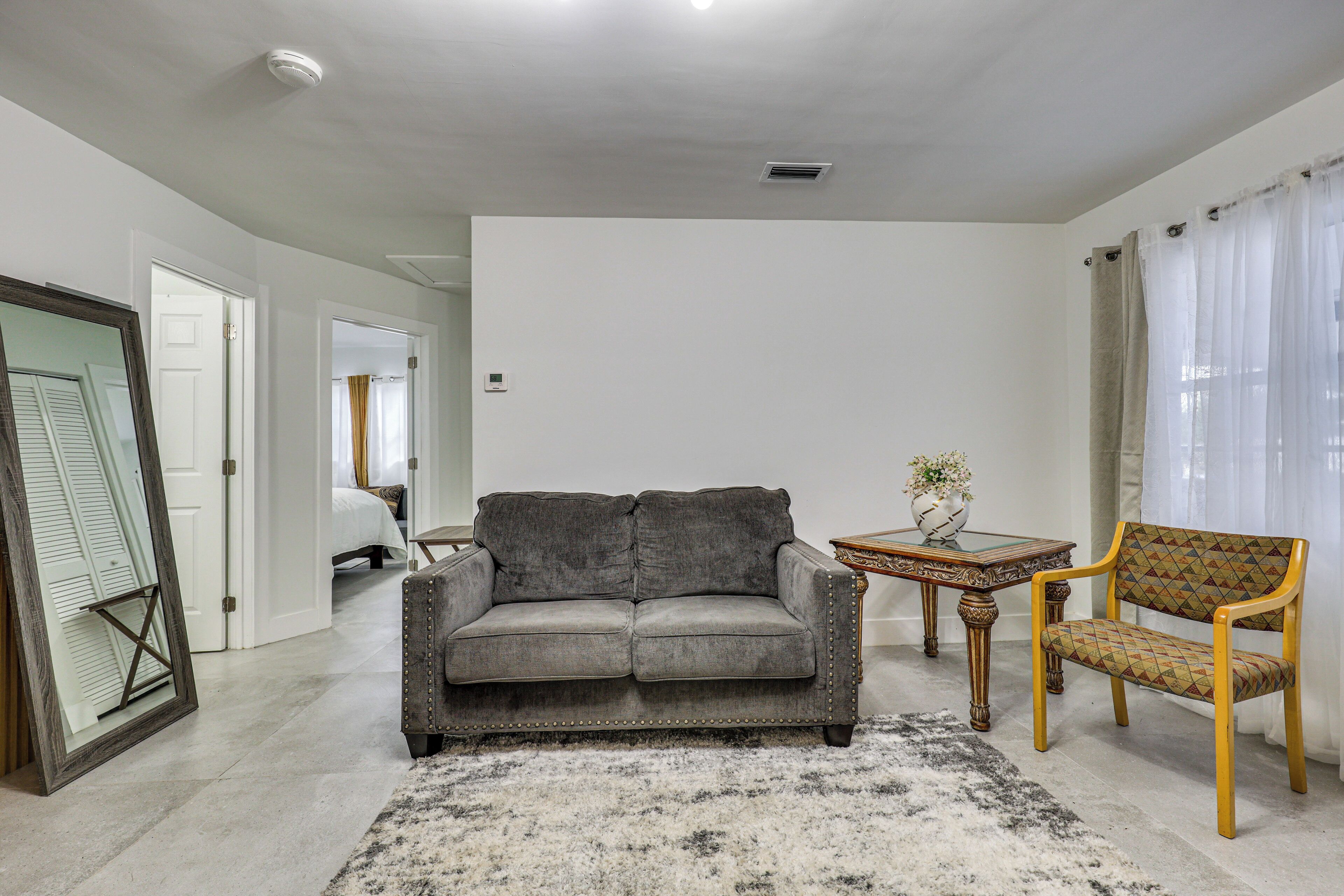 Una sala de estar con un sofá gris, un sillón estampado, una mesita con un jarrón y un espejo grande.