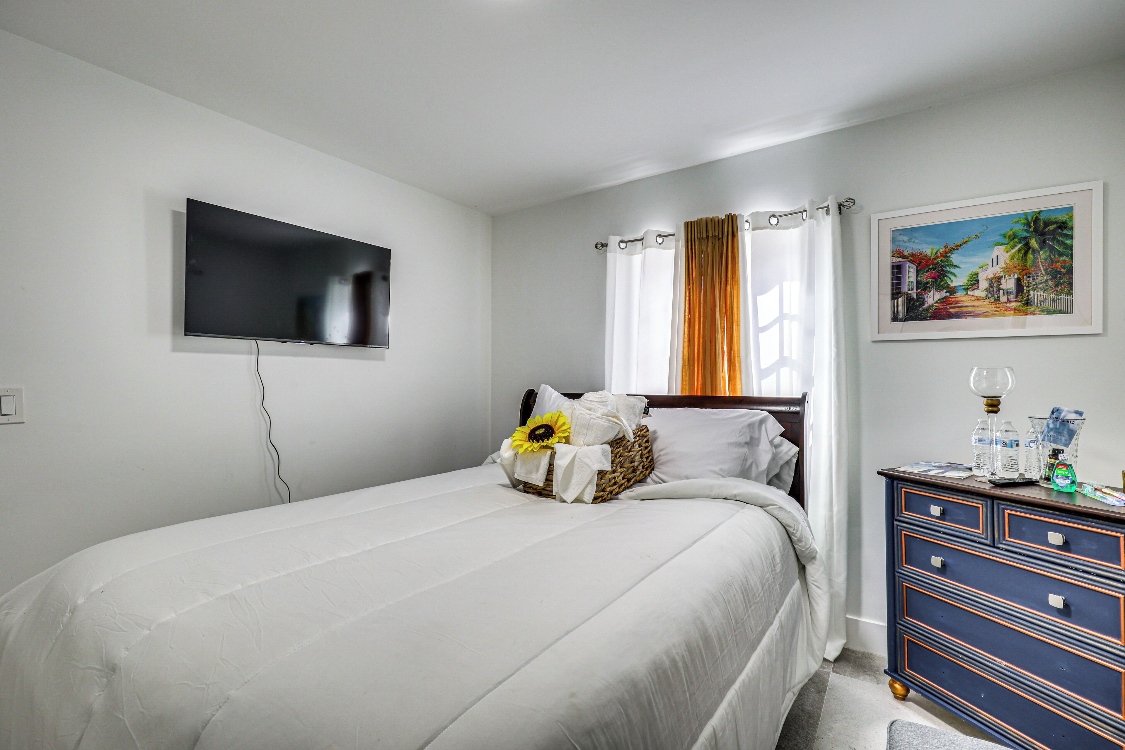 A bedroom with a bed, a television mounted on the wall, a dresser with drawers, and a painting on the wall.
