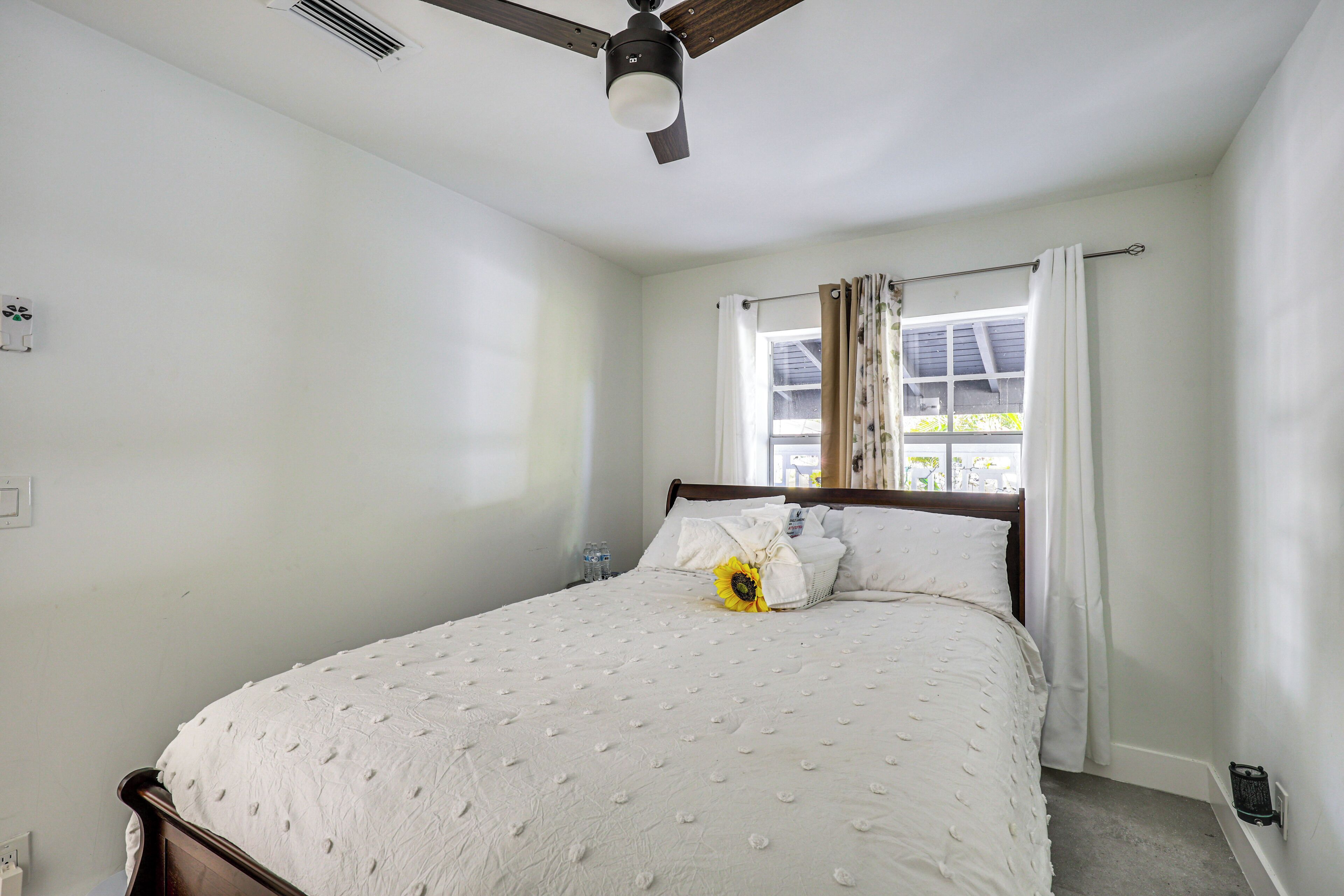 Ein Schlafzimmer mit Bett, Deckenventilator, Fenster mit Vorhängen und Nachttisch mit Lampe.