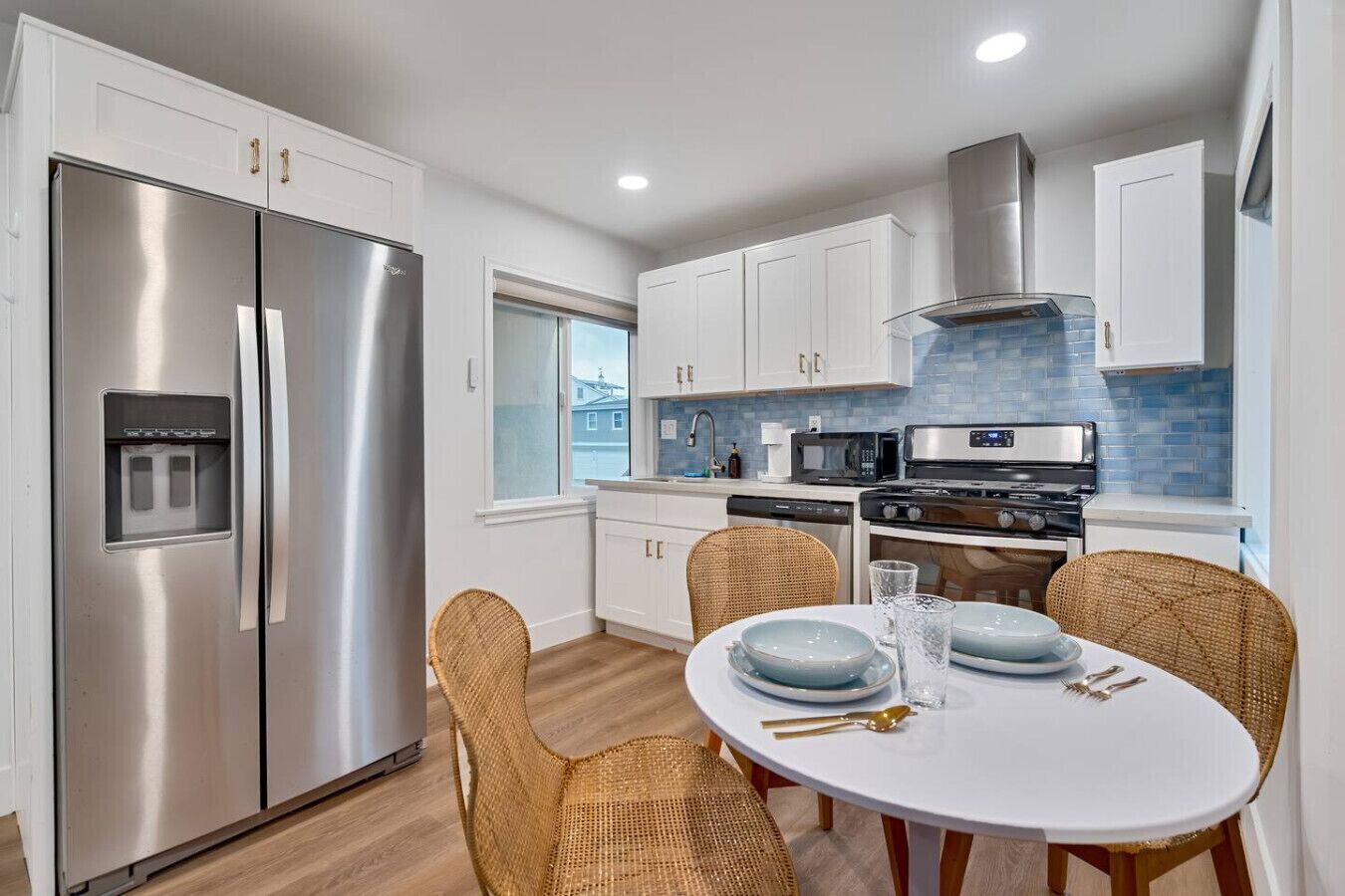 A modern kitchen with stainless steel appliances, white cabinetry, and a dining area with wicker chairs and a white table.
