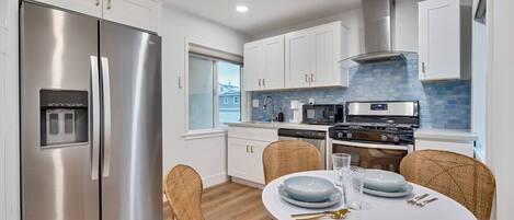 A modern kitchen with stainless steel appliances, white cabinetry, and a dining area with wicker chairs and a white table.