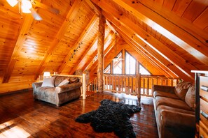 A log cabin interior with a wooden ceiling, a sofa, a rug, and a view through a window. - Serenity Now | Hocking Hills, Covered Hot Tub (Logan)