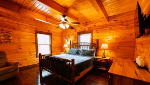 A wooden cabin bedroom with a bed, a chair, a nightstand, a lamp, and a ceiling fan.