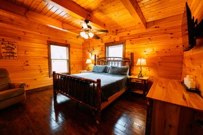 A wooden cabin bedroom with a bed, a chair, a nightstand, a lamp, and a ceiling fan.