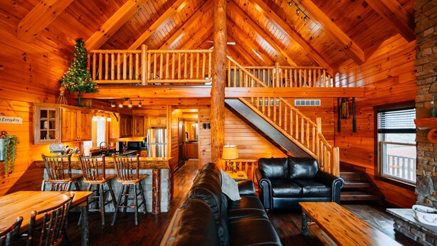 A log cabin interior with a dining area, a living room with a sofa and chairs, a staircase, and a Christmas tree.