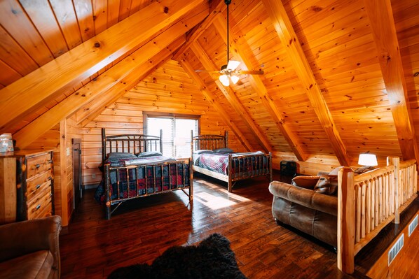 A log cabin with a high ceiling, wooden floors, two bunk beds, a sofa, and a ceiling fan.