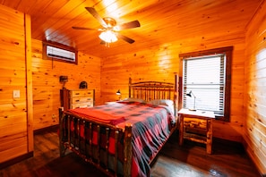 A wooden cabin bedroom with a bed, a desk, a ceiling fan, and a window.