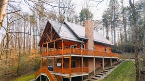 A two-story log cabin with a balcony, a stone chimney, and a staircase leading to the upper level.