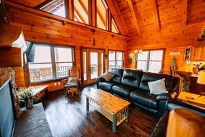 A log cabin interior with a black leather sofa, wooden coffee table, and large windows.