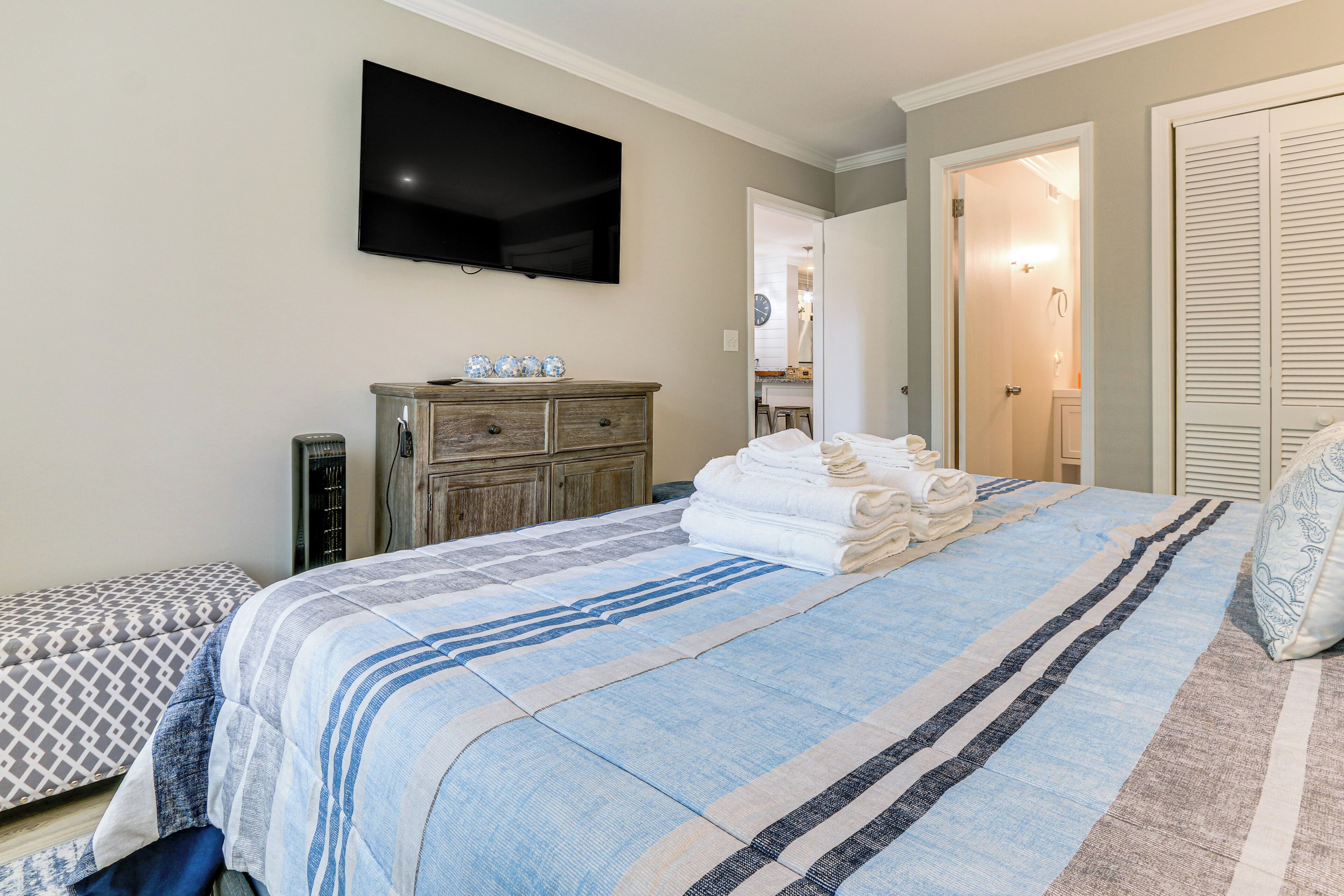A bedroom with a bed, a TV mounted on the wall, a wooden dresser, and a door leading to another room.