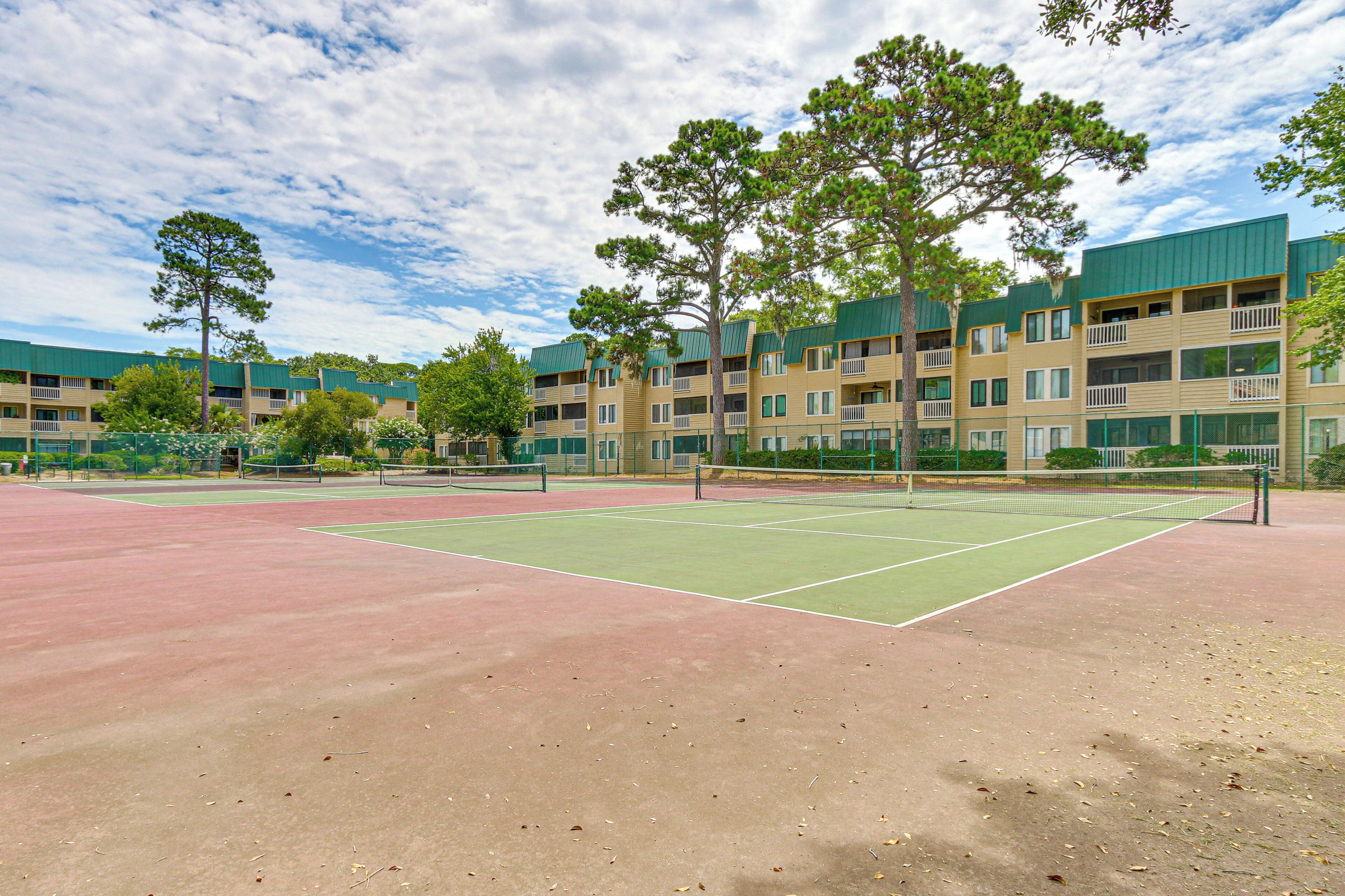 A tennis court with a net, surrounded by trees and a multi-story building with balconies.