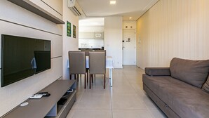 Ein Wohnzimmer mit Flachbildfernseher, einer Couch, einem Esstisch mit Stühlen und einer Küchenzeile im Hintergrund.