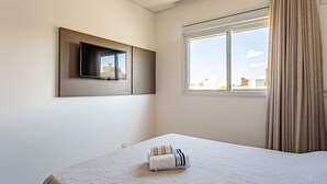 Ein Hotelzimmer mit Bett, einem an der Wand montierten Fernseher, einem Fenster mit Vorhängen und Blick auf die Stadt.