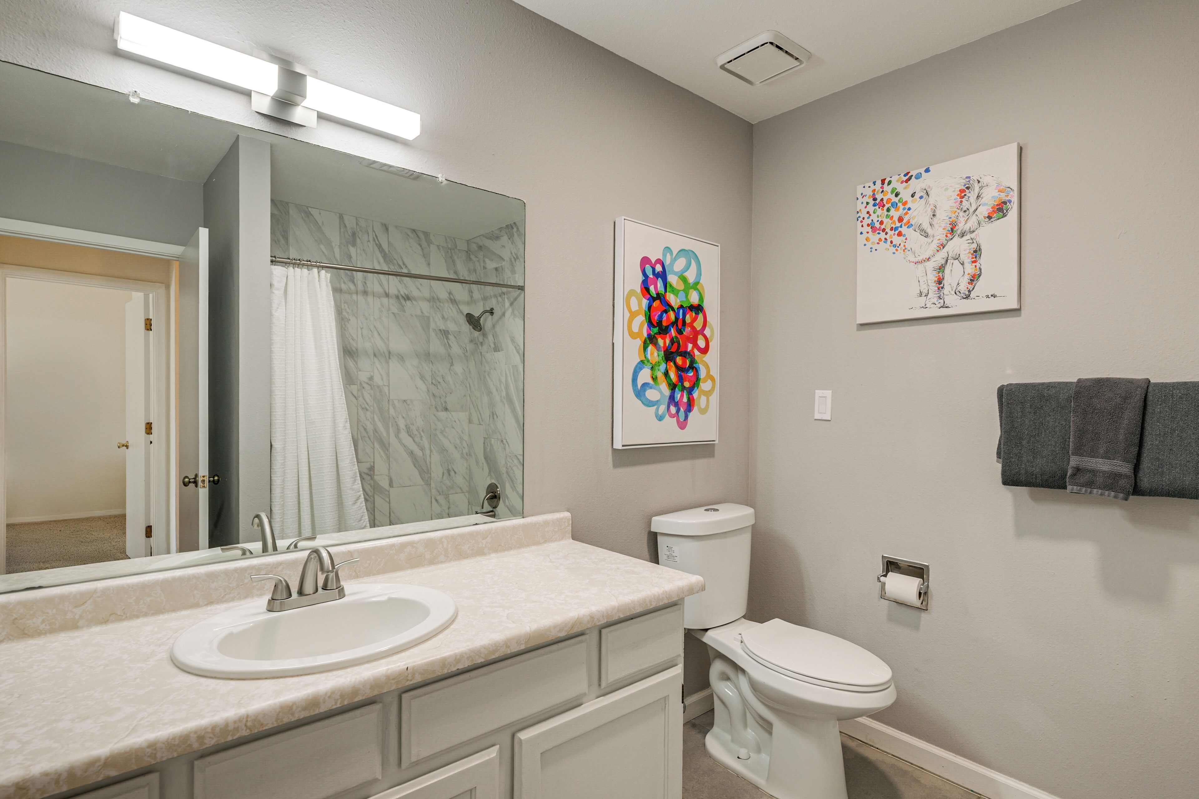 A bathroom with a marble sink, a toilet, a shower, and two wall-mounted artworks.