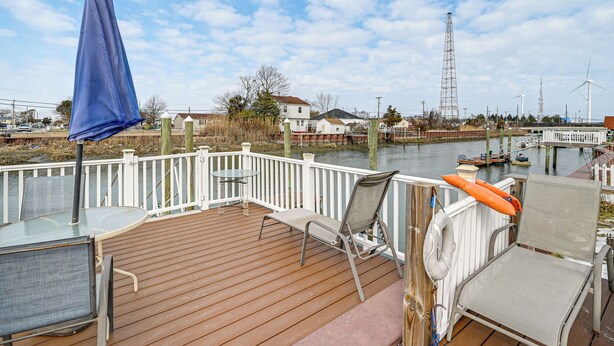Een houten terras met een glazen tafel en stoelen, uitkijkend op een wateroppervlak met boten en windturbines op de achtergrond.