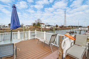 Une terrasse en bois avec une table et des chaises en verre, donnant sur une étendue d’eau où l’on aperçoit des bateaux et des éoliennes au loin.