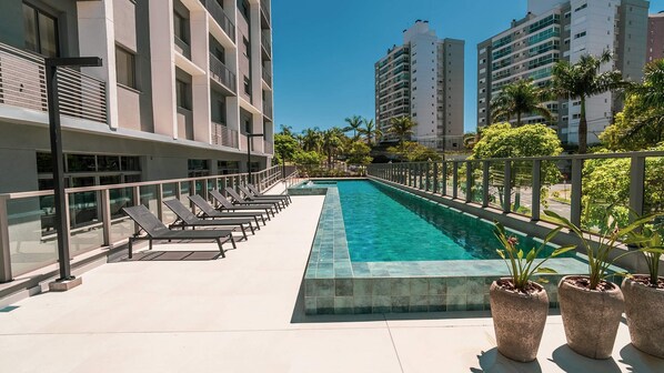 Una piscina en la azotea con sillones de descanso, cielo despejado y edificios altos circundantes.